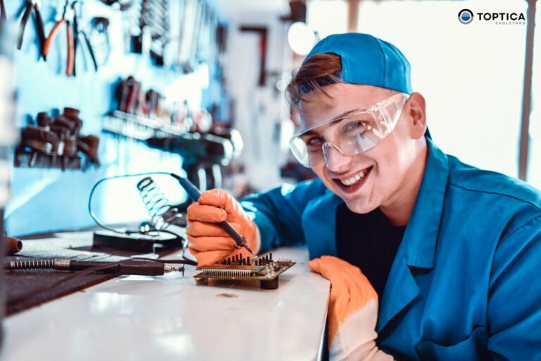 Ein Student der Fachrichtung Prozessentwicklung hantiert in einer Werkstatt für Fertigungstechnik fleißig mit einem Lötkolben an elektronischen Bauteilen, Schutzbrille, Handschuhe und blaue Mütze tragend.